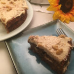 A slice of coconut carrot cake plated on a blue plate, a fork slightly hidden resting beside it with a white plate containing the remainder of the cake in the top left corner and a vivid yellow sunflower in the top right corner