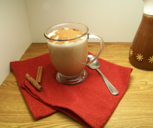 Gingerbread Coffee Syrup in an old fashioned syrup dispenser with a mug of gingerbread coffee in a clear mug with a spoon on one side and cinnamon sticks on the other, a red cloth underneath