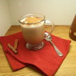 Sugar Free Gingerbread Coffee Syrup in an old fashioned dispenser to the back right with a mug of gingerbread coffee in a clear handled mug with a spoon on one side and cinnamon sticks on the other on top of a red cloth on a wooden surface