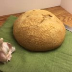 Garlic Parmesan Sourdough Bread whole and uncut, sitting on a green placemat with a bread knife beside it and a partial bulb of garlic to the front