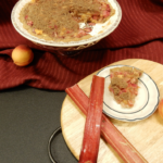 Peach Rhubarb Pie from an overhead angle, a slice of pie on a blue and white dessert plate with rhubarb to the left, an elevated pie dish with the remainder of the pie to the back on a red cloth