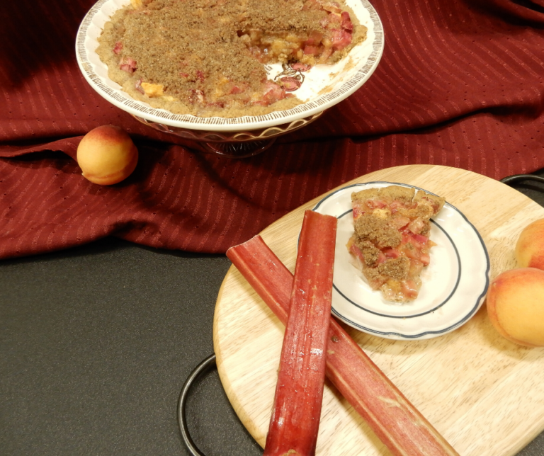 Peach Rhubarb Pie from an overhead angle, a slice of pie on a blue and white dessert plate with rhubarb to the left, an elevated pie dish with the remainder of the pie to the back on a red cloth