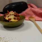 Stuffed Eggplant in a homemade green ceramic bowl with a wooden spoon to the right and a checkered red and white cloth behind with a whole eggplant on top